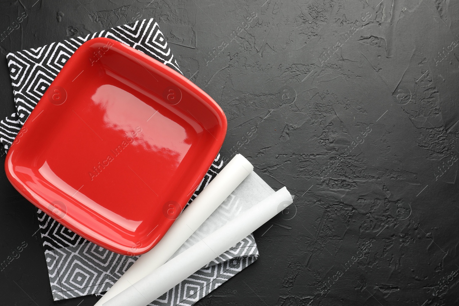 Photo of Red ceramic casserole and baking parchment paper on black table, top view. Space for text