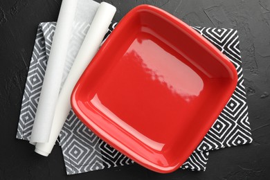 Photo of Red ceramic casserole and baking parchment paper on black table, top view