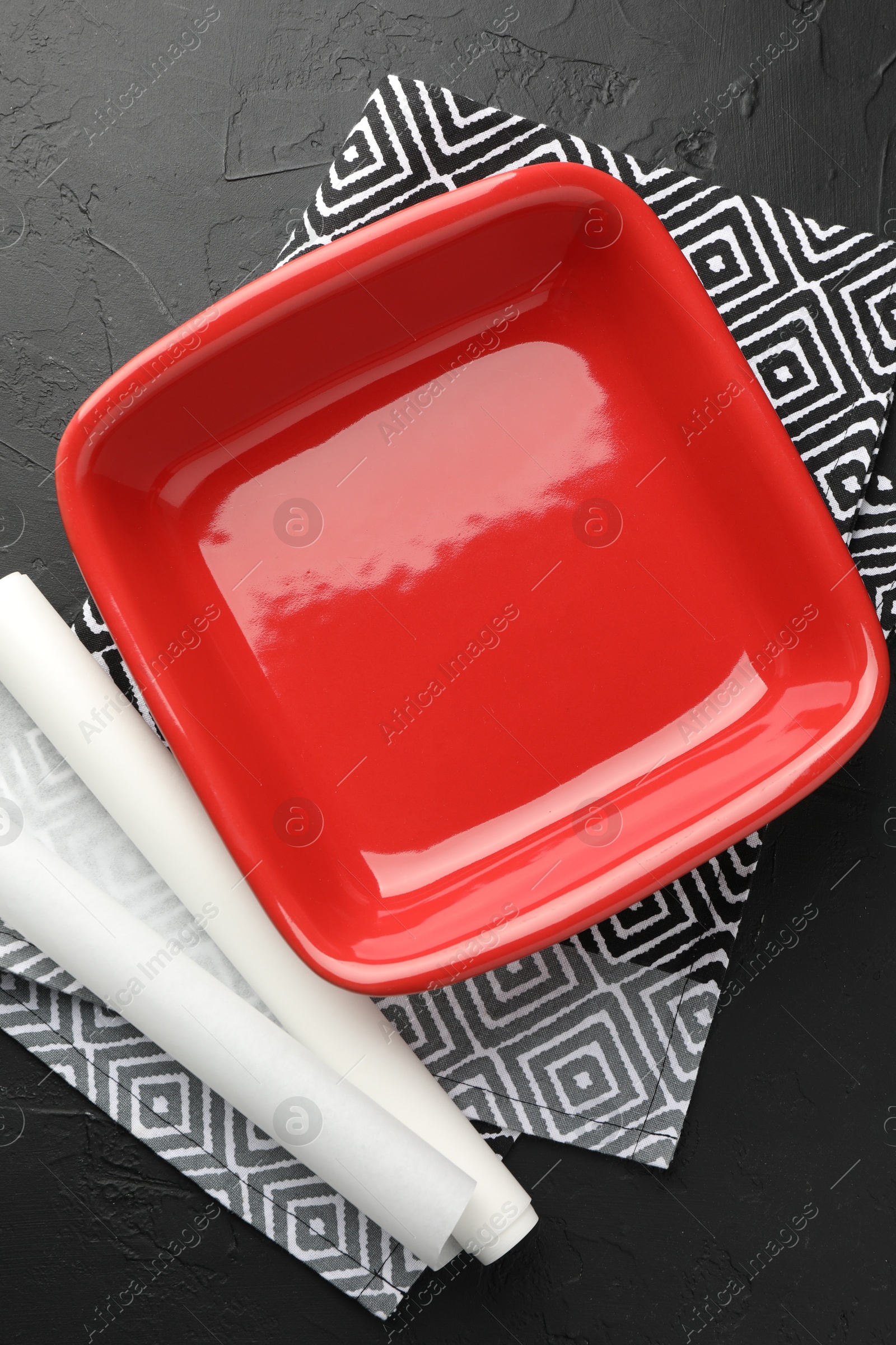 Photo of Red ceramic casserole and baking parchment paper on black table, top view