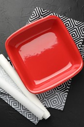 Photo of Red ceramic casserole and baking parchment paper on black table, top view