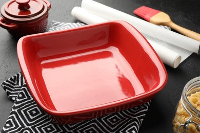 Photo of Red ceramic casseroles, baking parchment paper, raw pasta and brush on black table, closeup