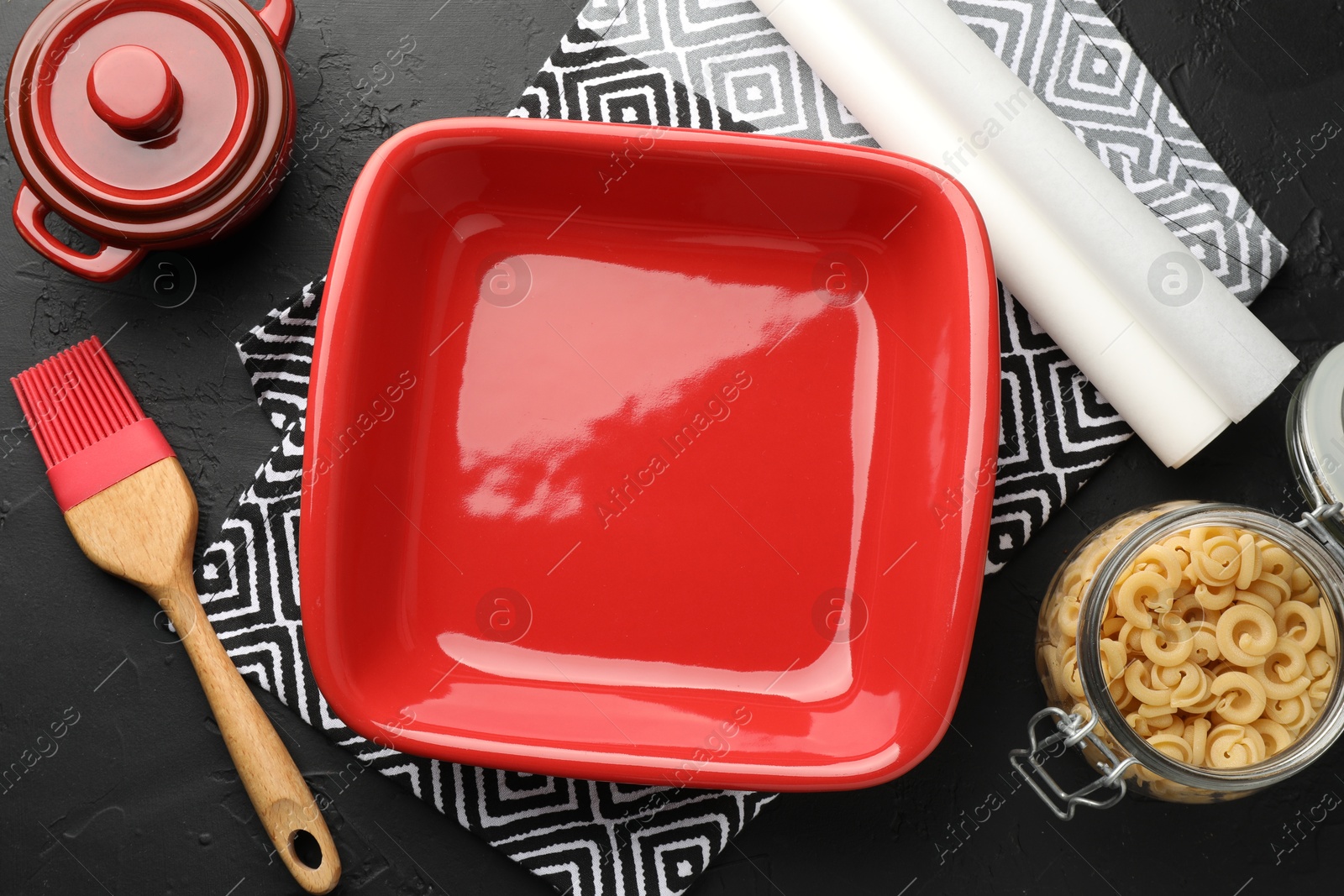 Photo of Red ceramic casseroles, baking parchment paper, raw pasta and brush on black table, flat lay