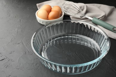 Photo of Glass casserole, whisk and eggs on black table, closeup. Space for text