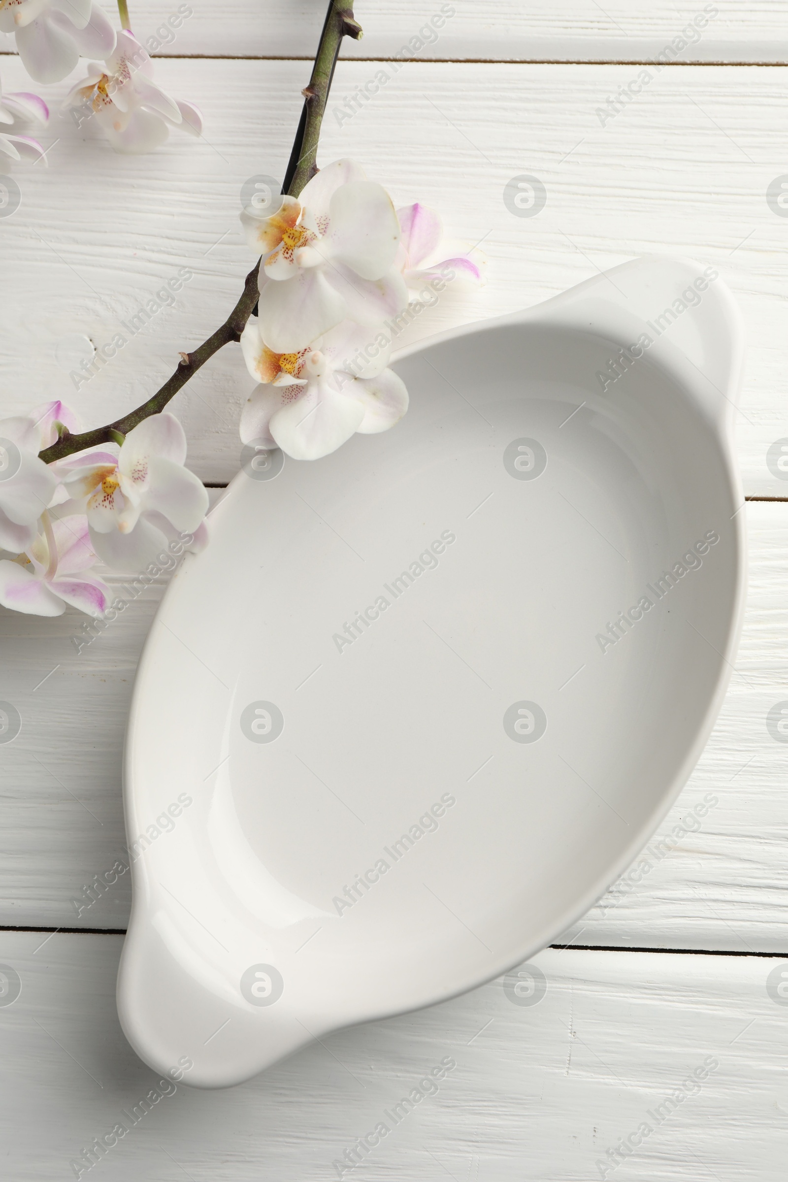 Photo of Ceramic casserole and orchid branch on white wooden table, top view