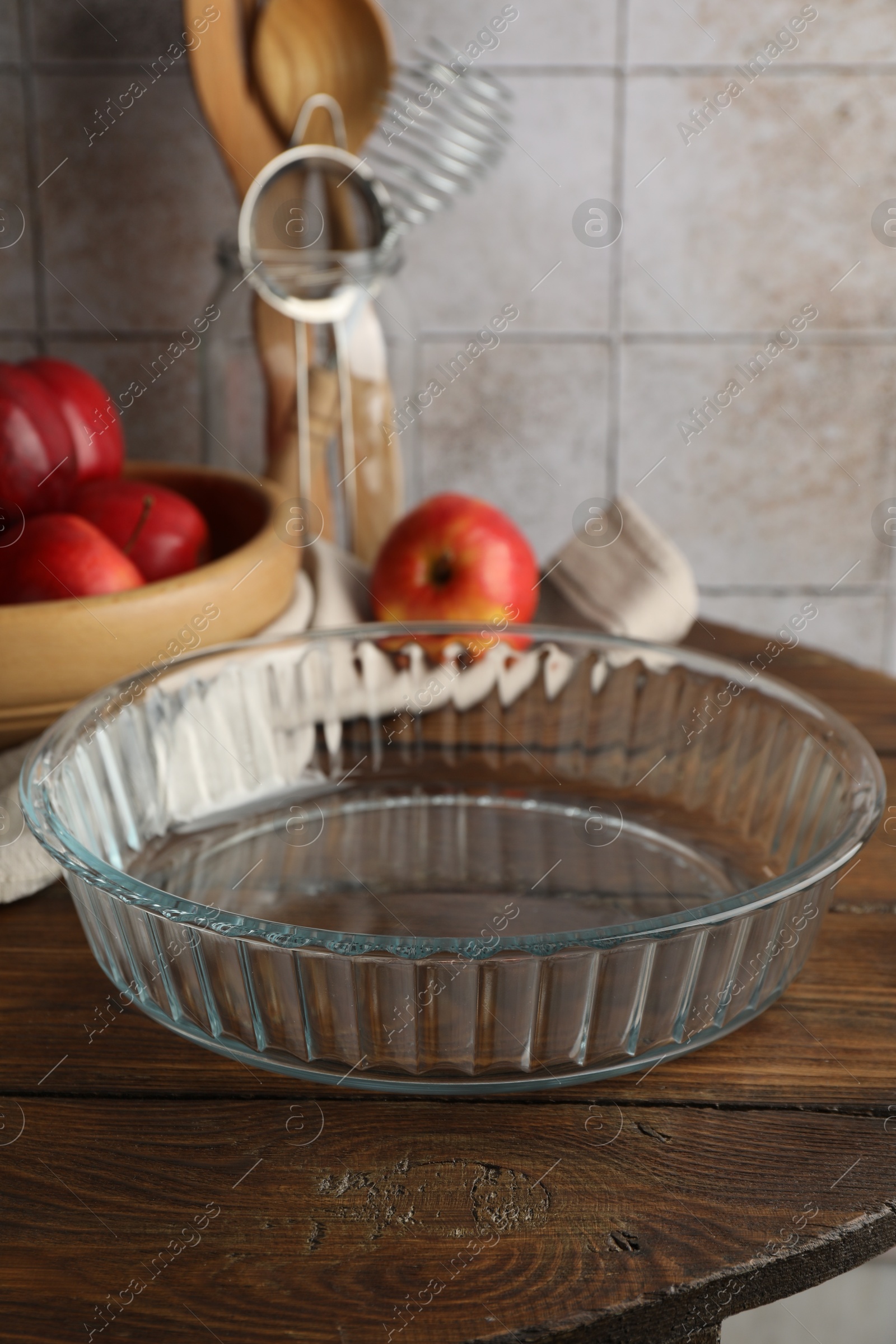 Photo of Glass casserole, apples and different cooking utensils on wooden table, closeup