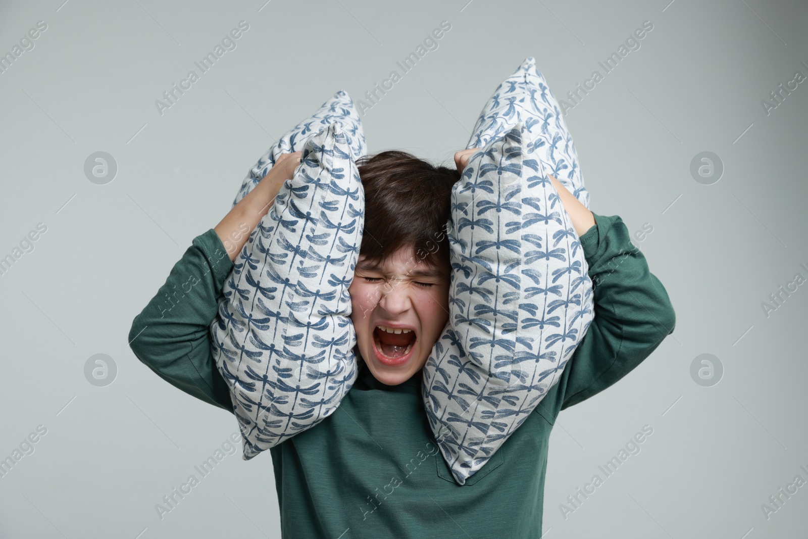 Photo of Annoyed boy covering his ears with pillows due to loud sound on grey background