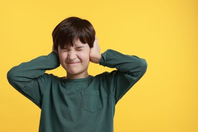 Annoyed boy covering his ears due to loud sound on yellow background, space for text