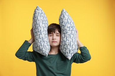 Photo of Annoyed boy covering his ears with pillows due to loud sound on yellow background