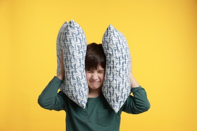 Photo of Annoyed boy covering his ears with pillows due to loud sound on yellow background