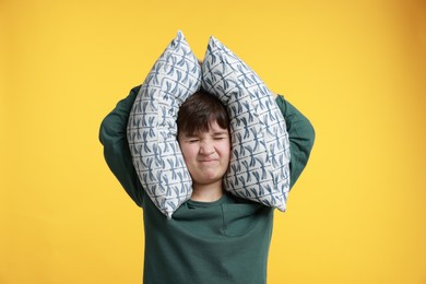 Photo of Annoyed boy covering his ears with pillows due to loud sound on yellow background