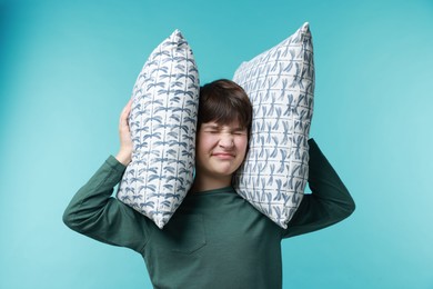 Photo of Annoyed boy covering his ears with pillows due to loud sound on light blue background