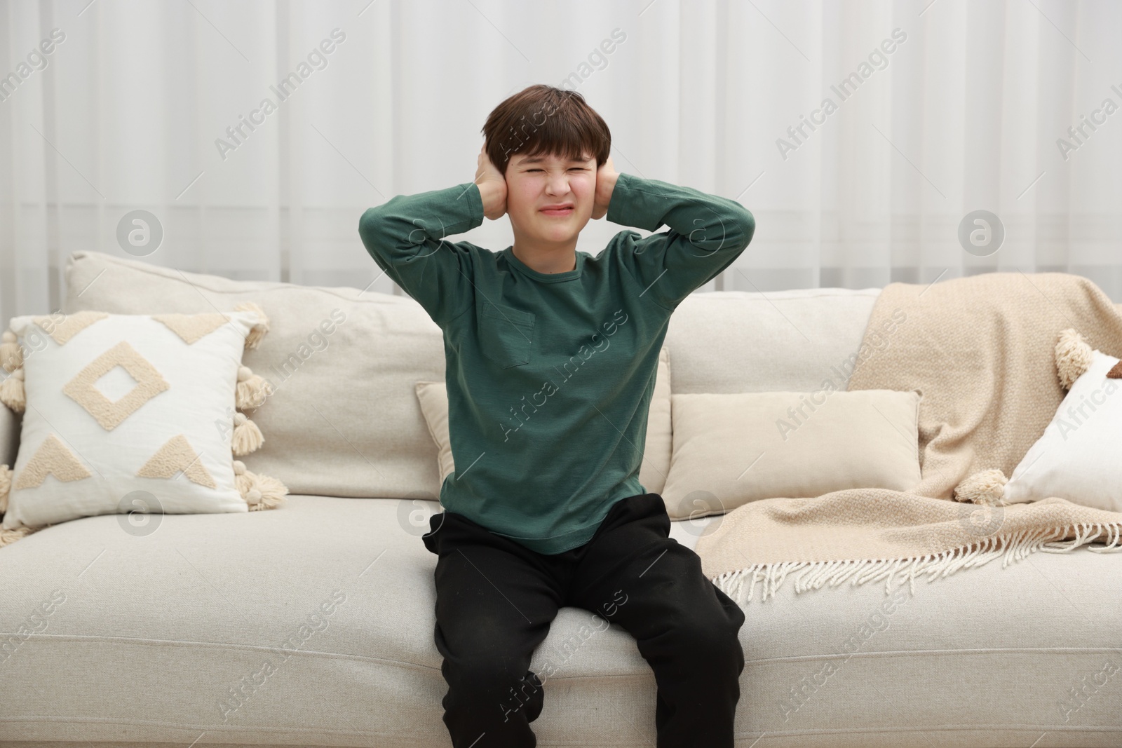 Photo of Annoyed boy covering his ears due to loud sound on sofa at home