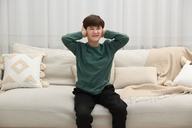 Photo of Annoyed boy covering his ears due to loud sound on sofa at home