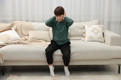 Photo of Annoyed boy covering his ears due to loud sound on sofa at home