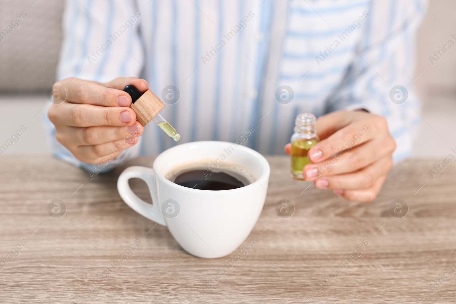 Photo of Young woman putting CBD tincture into cup with drink at wooden table, closeup