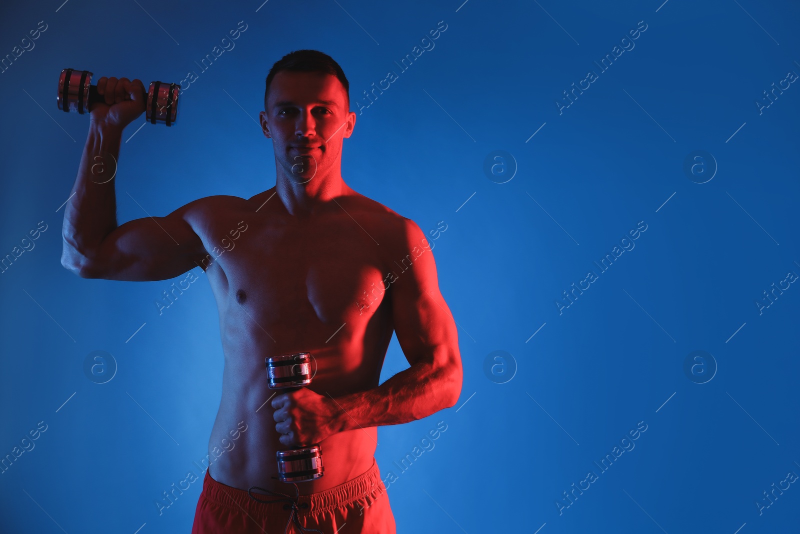 Photo of Man exercising with dumbbells in red light on blue background, space for text