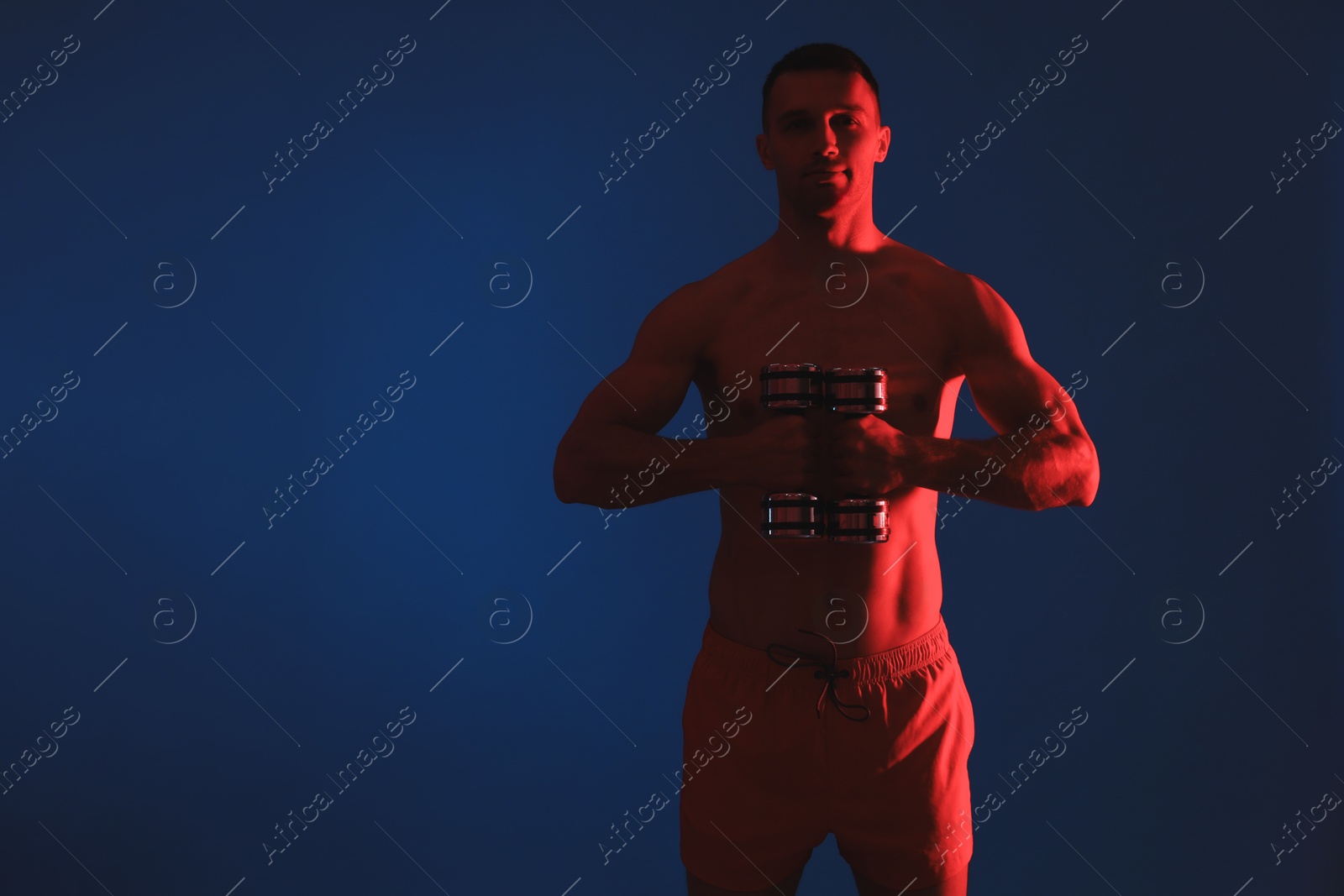 Photo of Man exercising with dumbbells in red light on blue background. Space for text