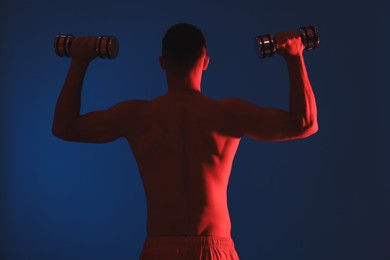 Photo of Man exercising with dumbbells in red light on blue background, back view