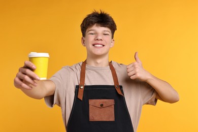 Photo of Teenage boy with cup of coffee working as barista on orange background