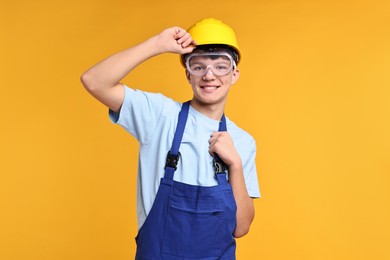 Photo of Teenage boy in hardhat and protective mask working as builder on orange background