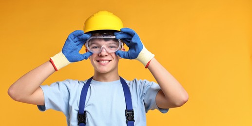 Teenage boy in hardhat and protective mask working as builder on orange background