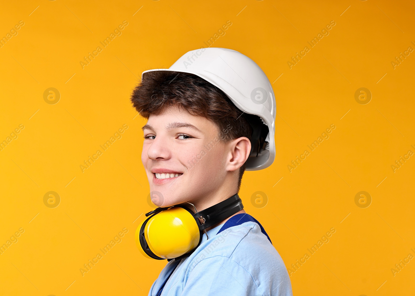 Photo of Teenage boy in hardhat with earmuffs working as builder on orange background