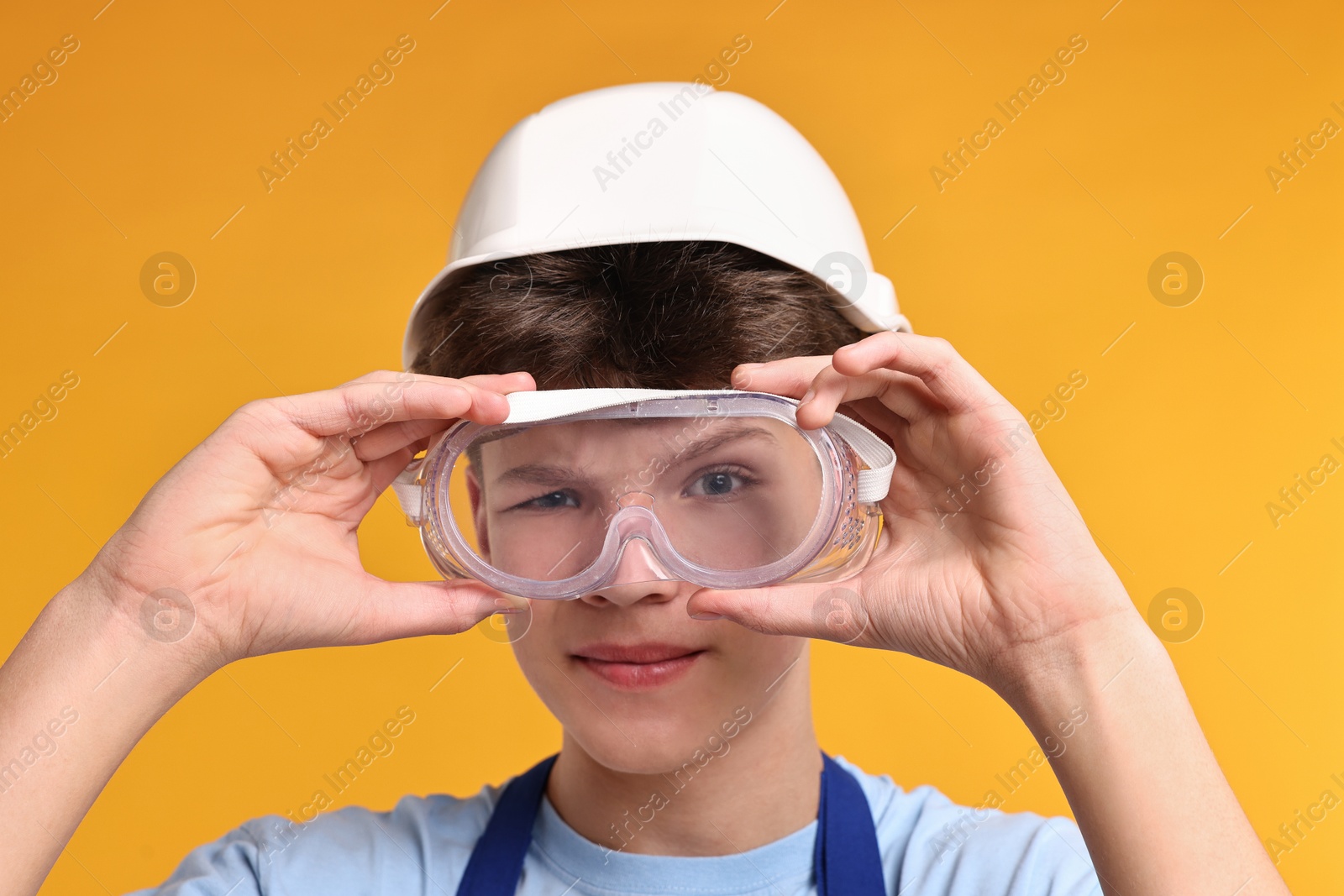 Photo of Teenage boy in hardhat and protective mask working as builder on orange background