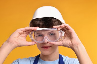 Photo of Teenage boy in hardhat and protective mask working as builder on orange background