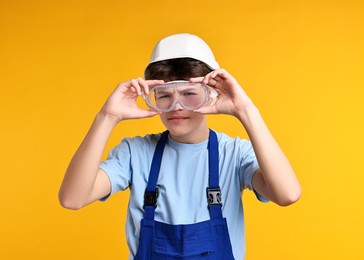 Photo of Teenage boy in hardhat and protective mask working as builder on orange background