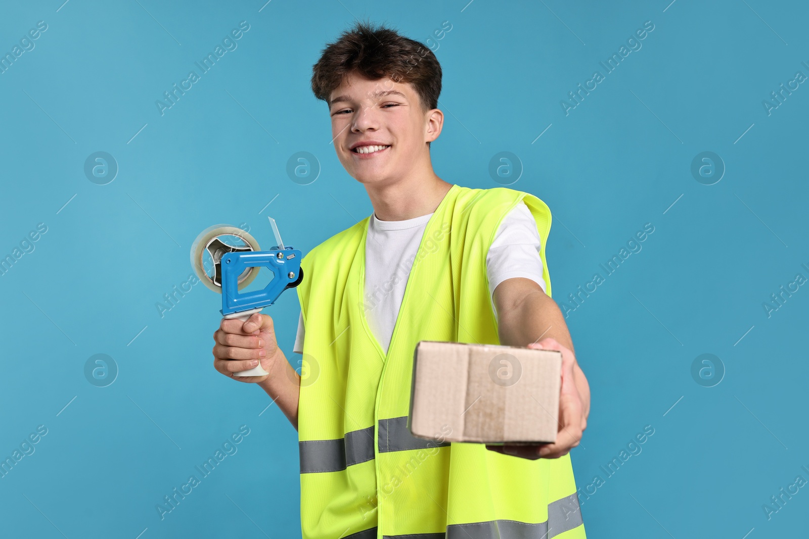 Photo of Teenage boy with tape gun dispenser and box in safety vest working as warehouse packer on blue background