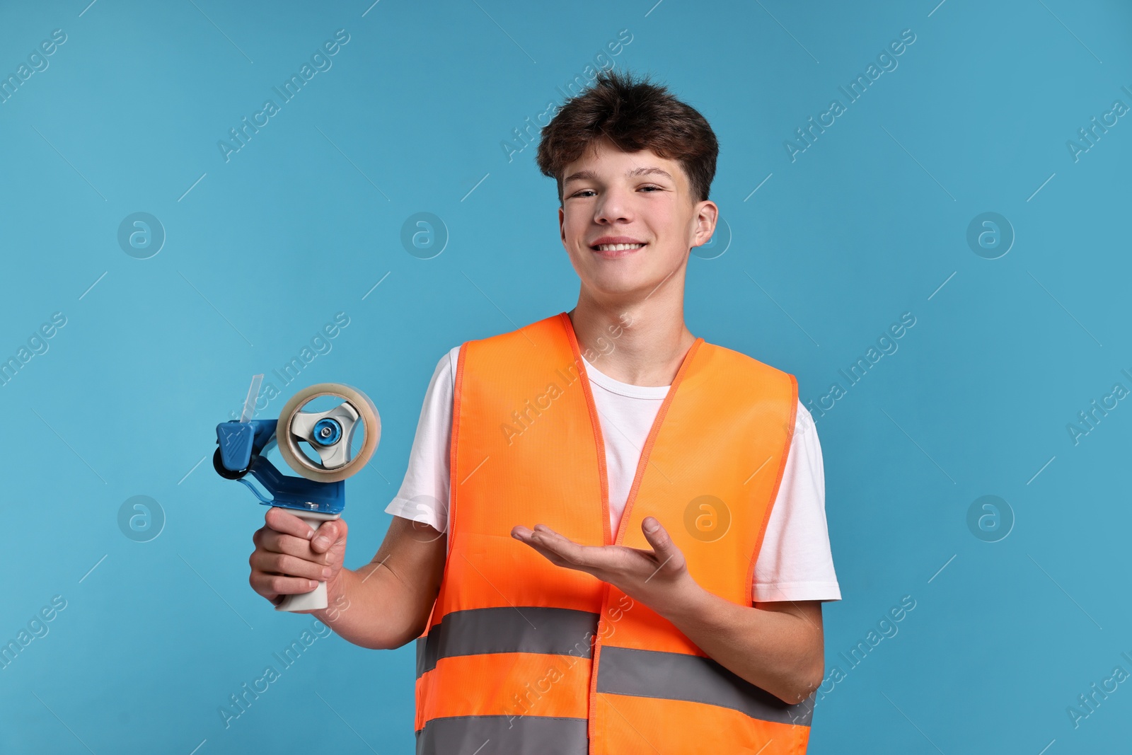 Photo of Teenage boy with tape gun dispenser in safety vest working as warehouse packer on blue background