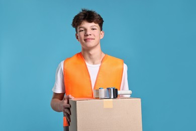 Photo of Teenage boy with tape gun dispenser and box in safety vest working as warehouse packer on blue background