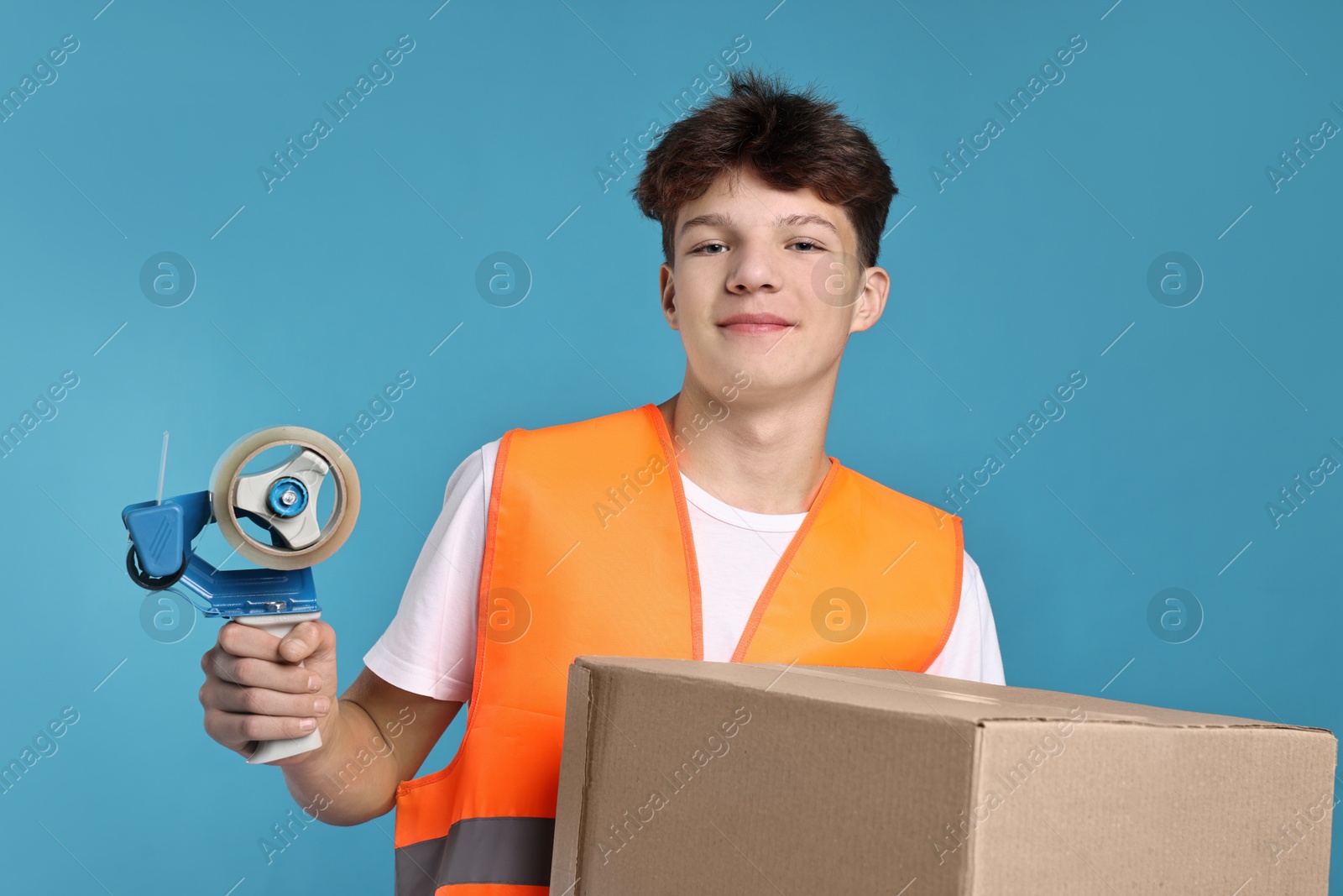 Photo of Teenage boy with tape gun dispenser and box in safety vest working as warehouse packer on blue background