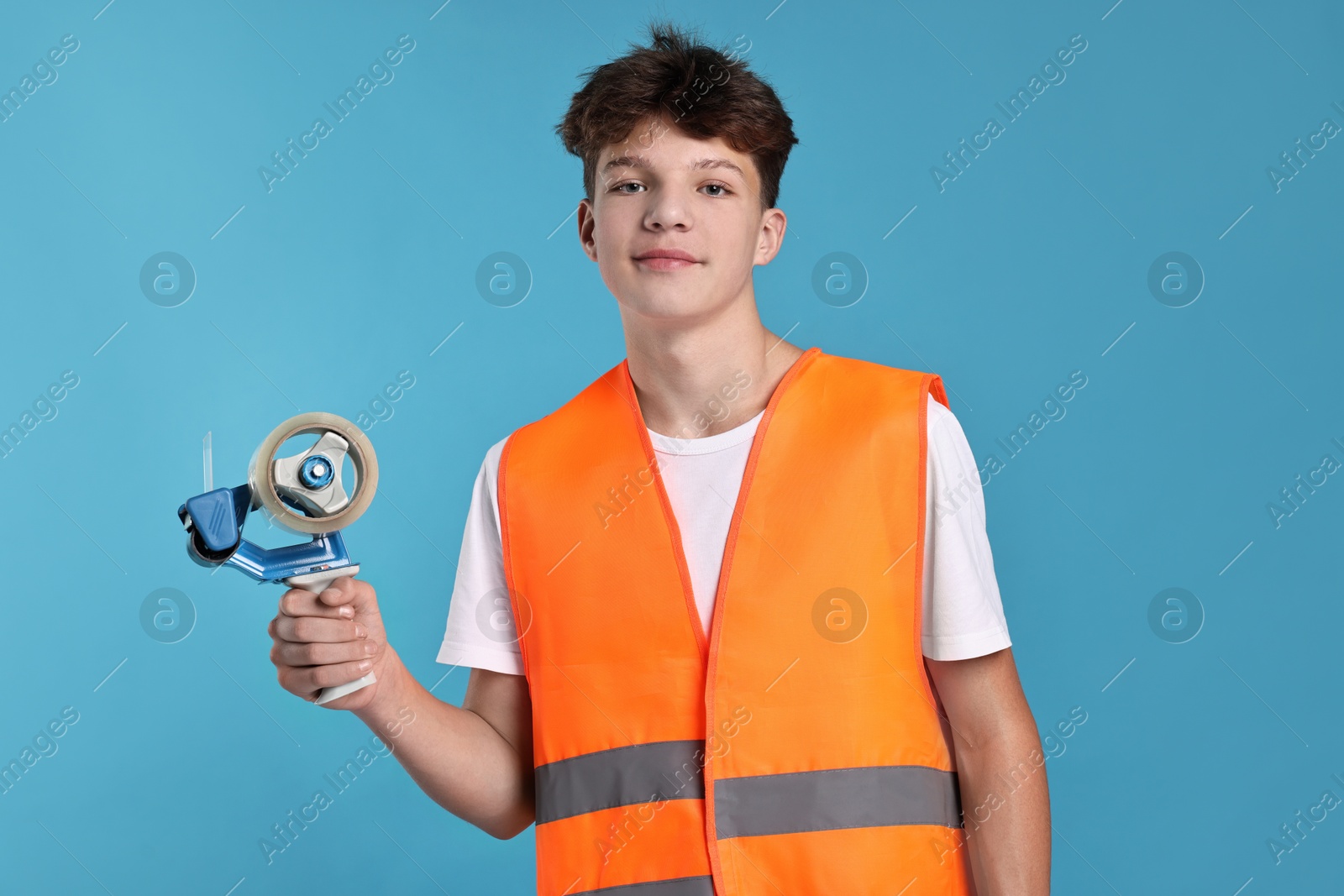 Photo of Teenage boy with tape gun dispenser in safety vest working as warehouse packer on blue background