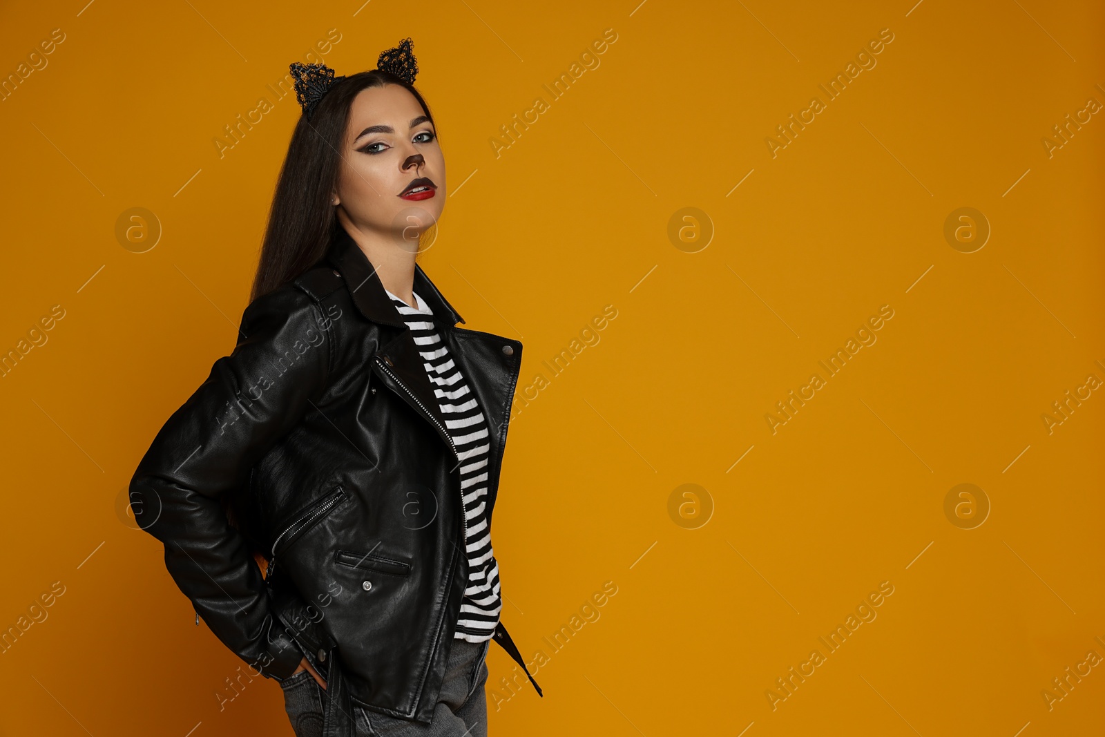 Photo of Woman with cat makeup and ears in leather jacket on orange background. Space for text