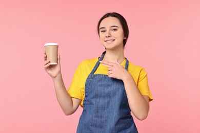 Photo of Girl in apron with paper cup of coffee on pink background. Work for teenagers