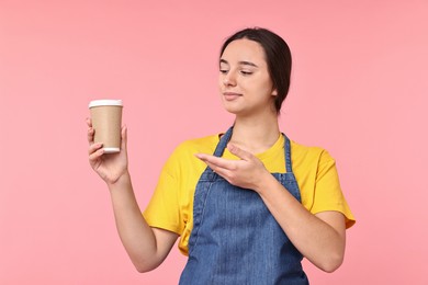 Photo of Girl in apron with paper cup of coffee on pink background. Work for teenagers