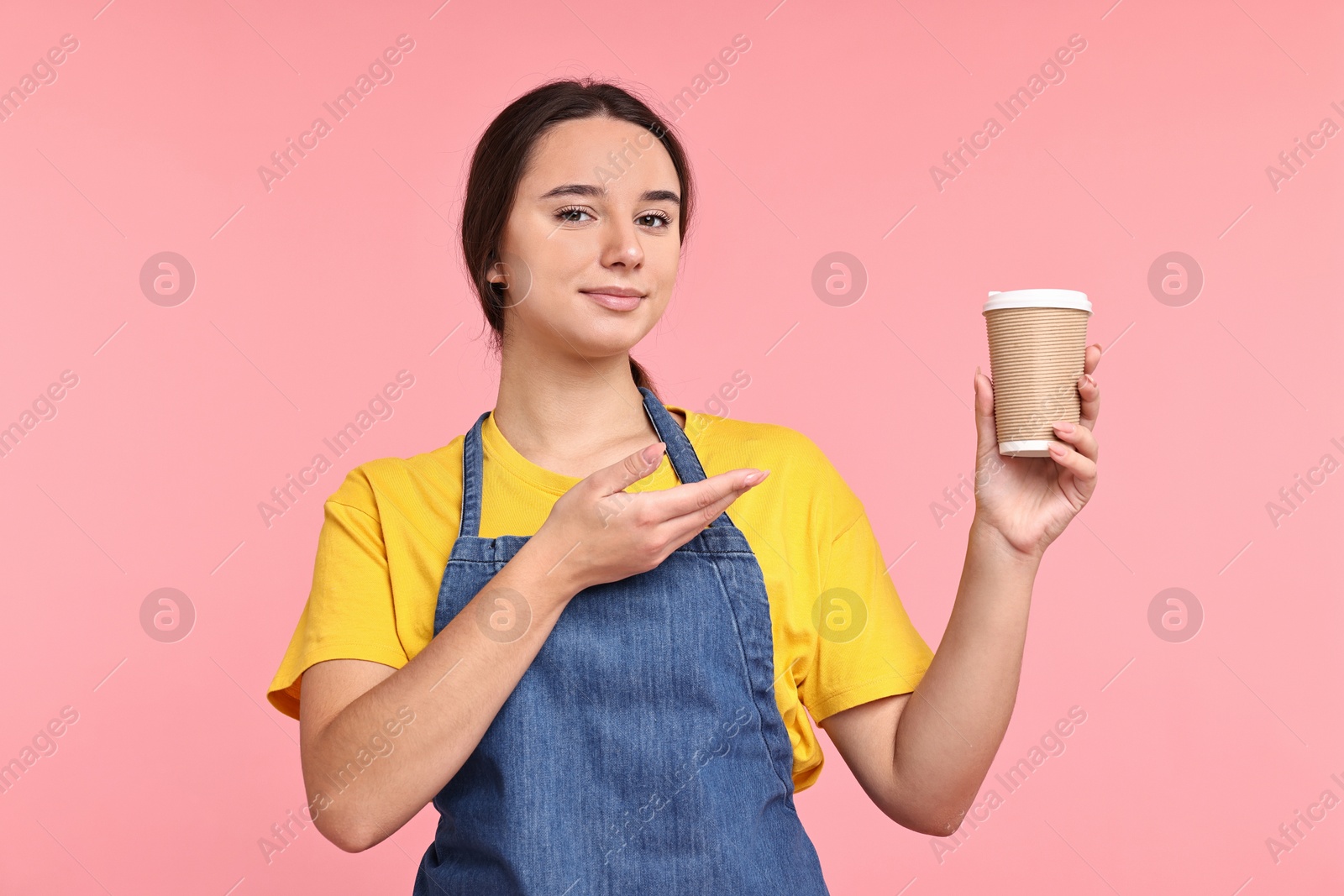 Photo of Girl in apron with paper cup of coffee on pink background. Work for teenagers