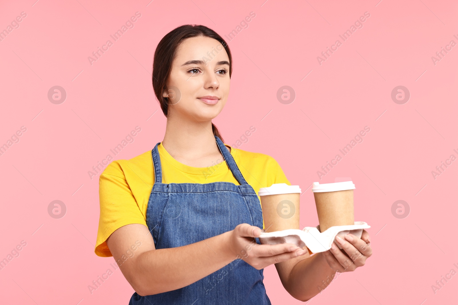 Photo of Girl in apron with takeaway paper cups of coffee on pink background. Work for teenagers