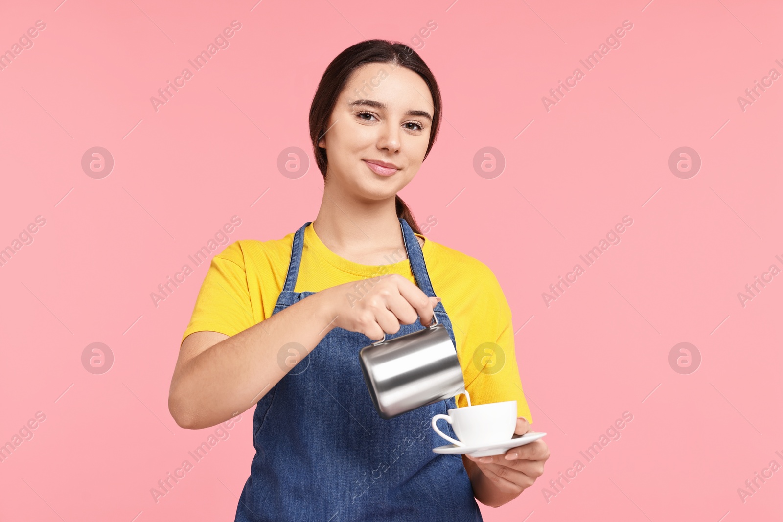 Photo of Girl in apron pouring milk from pitcher into cup of coffee on pink background. Work for teenagers