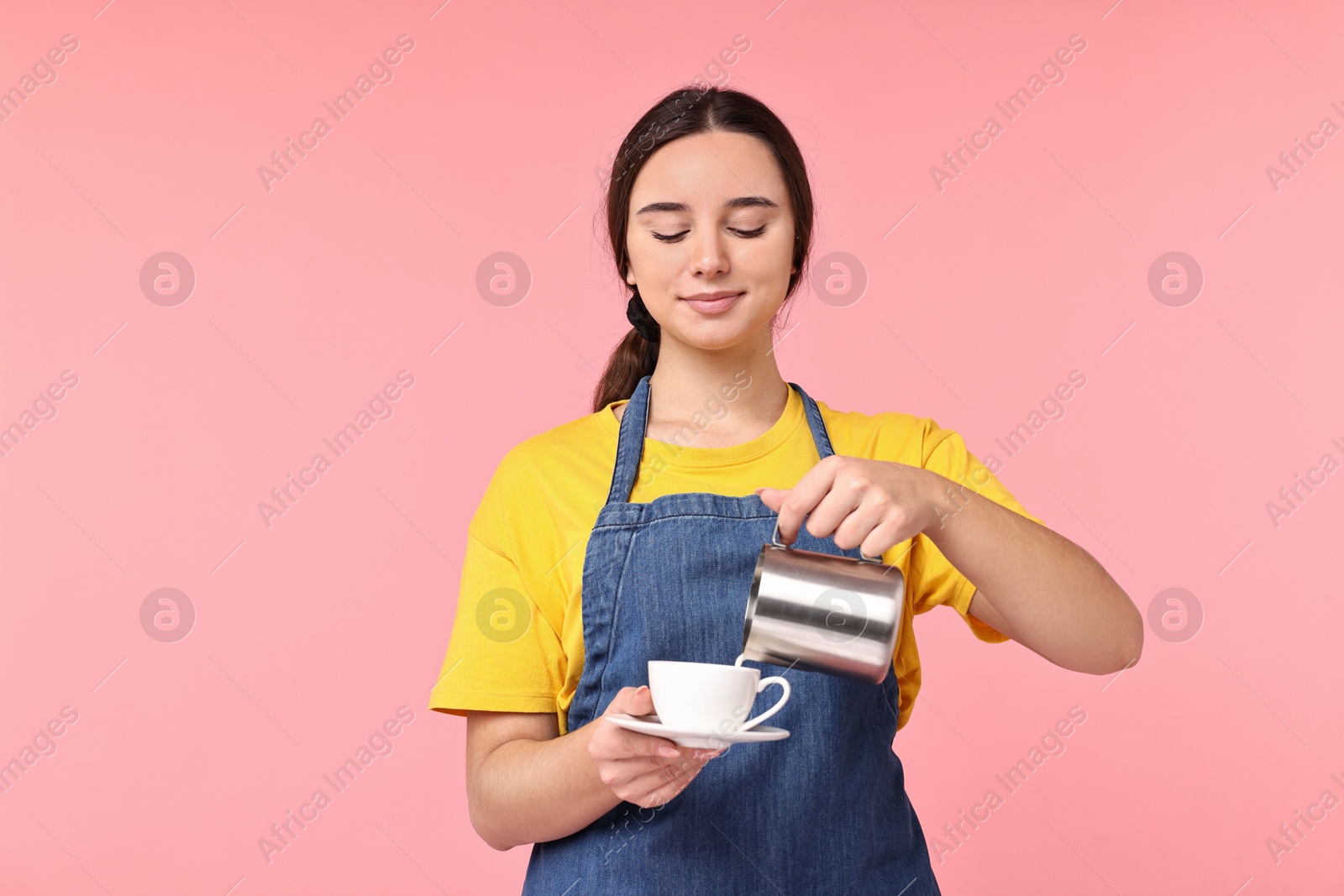 Photo of Girl in apron pouring milk from pitcher into cup of coffee on pink background. Work for teenagers