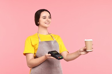 Photo of Girl in apron with paper cup of coffee and payment terminal on pink background. Work for teenagers