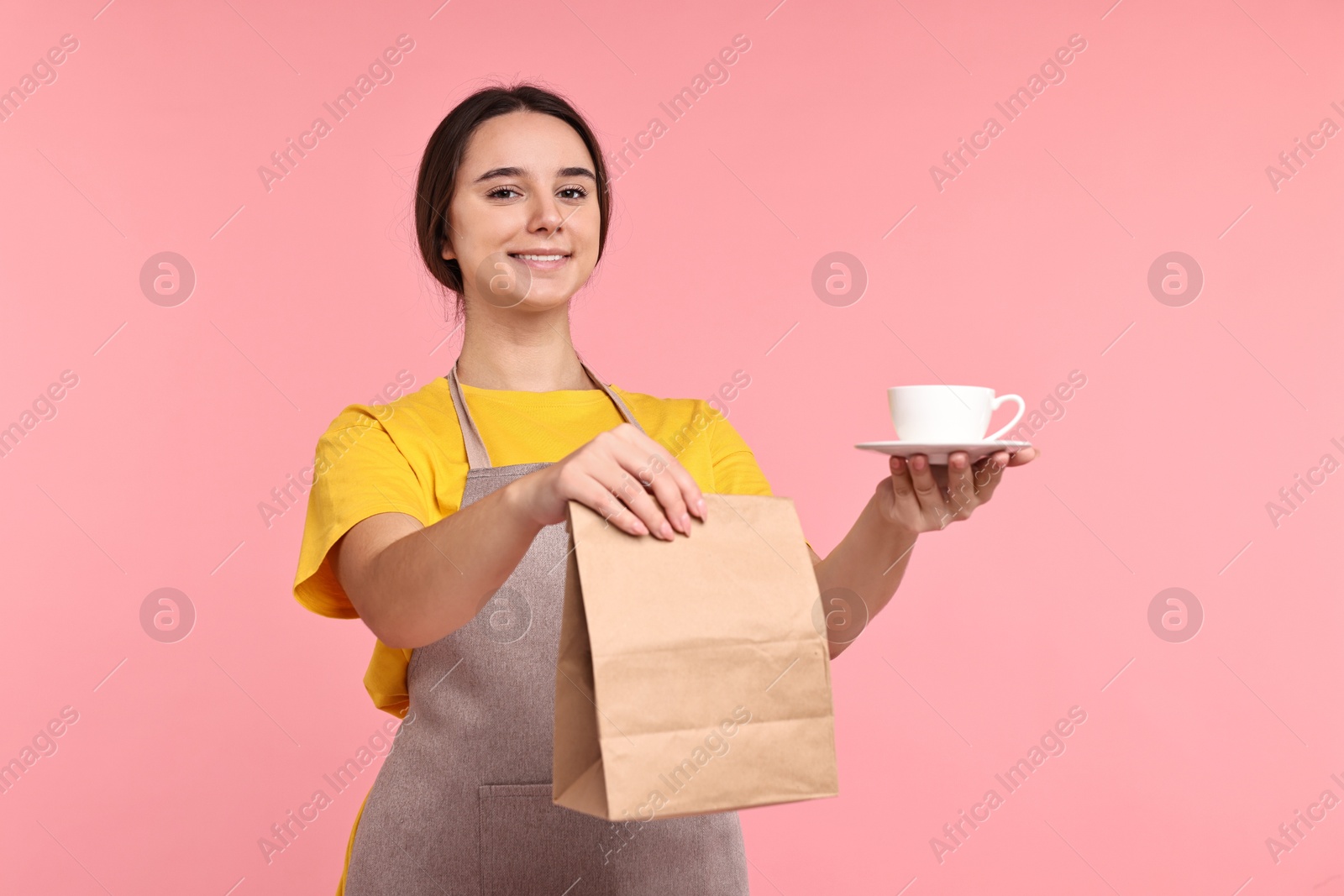 Photo of Girl in apron holding cup of coffee and paper bag on pink background. Work for teenagers