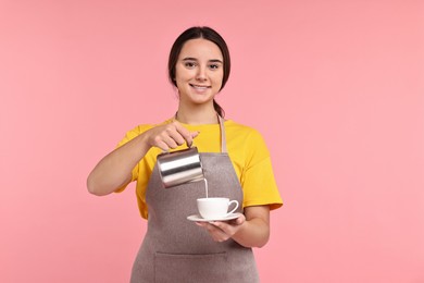 Photo of Girl in apron pouring milk from pitcher into cup of coffee on pink background. Work for teenagers