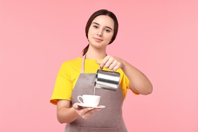 Photo of Girl in apron pouring milk from pitcher into cup of coffee on pink background. Work for teenagers