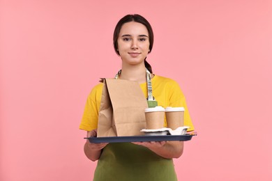 Girl in apron holding takeaway cups of coffee and paper bag on pink background. Work for teenagers