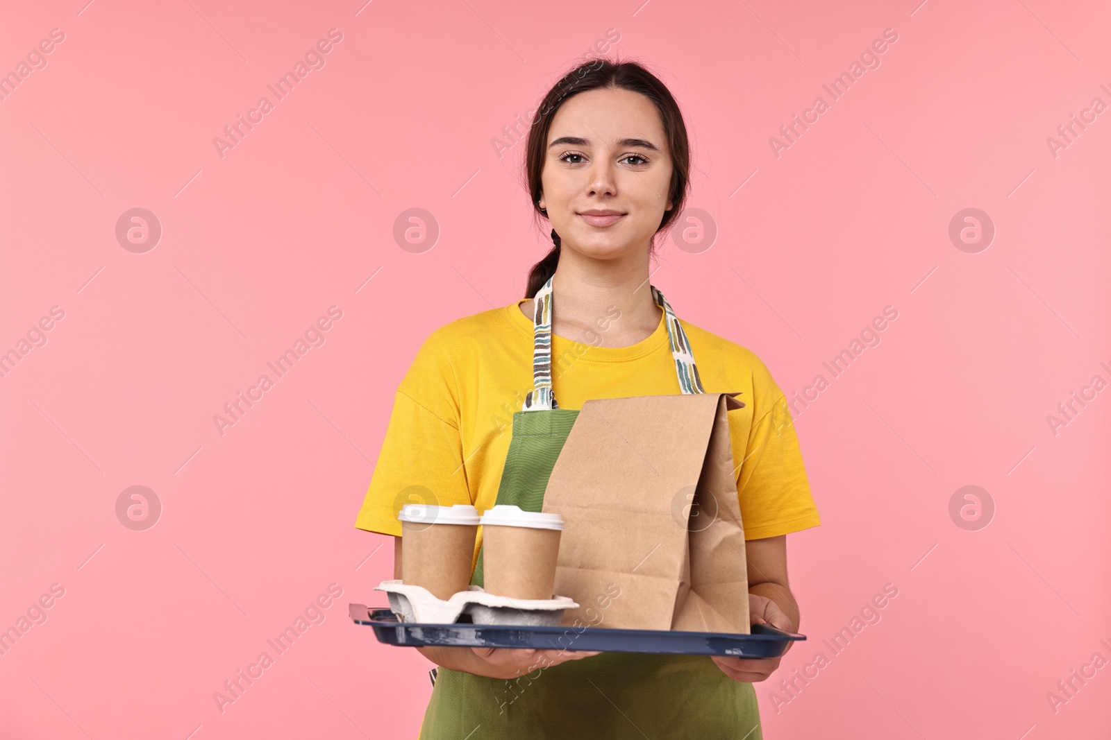 Photo of Girl in apron holding takeaway cups of coffee and paper bag on pink background. Work for teenagers