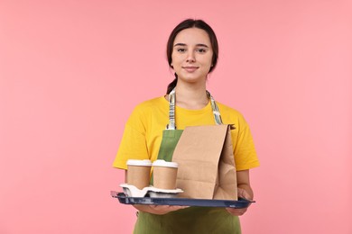 Photo of Girl in apron holding takeaway cups of coffee and paper bag on pink background. Work for teenagers