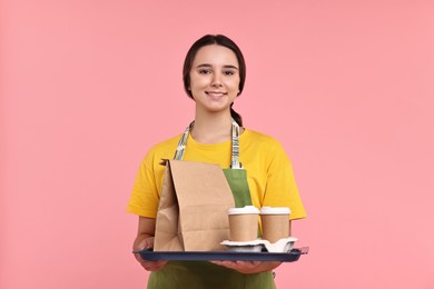 Photo of Girl in apron holding takeaway cups of coffee and paper bag on pink background. Work for teenagers