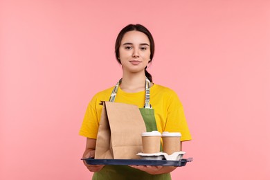 Photo of Girl in apron holding takeaway cups of coffee and paper bag on pink background. Work for teenagers